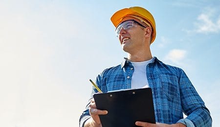 A man wearing a yellow hard hat, safety glasses, and a blue plaid shirt holds a clipboard and pencil while looking off to the side, ready for tree services Long Island under the bright blue NY sky.