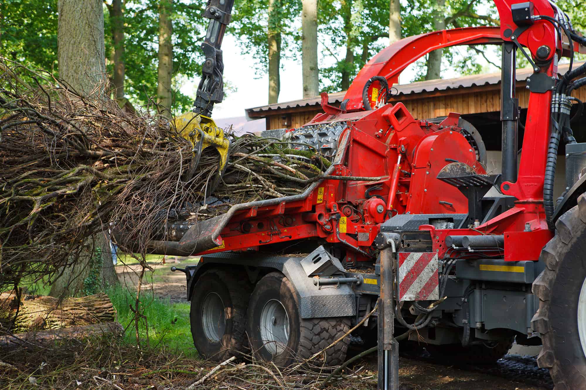 A wood chipper attached to a tractor processes a pile of tree branches and logs in a forested NY area near a wooden building, showcasing professional tree services Long Island relies on. The red machinery is actively pulling branches inside.
